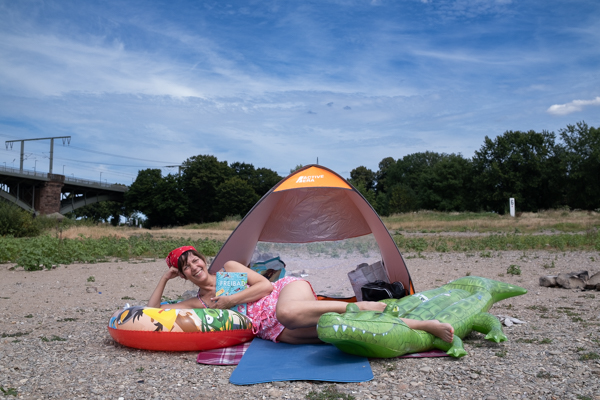 Arzu Ermen liegt vor einem kleinen Zelt auf Steinen, direkt am Rhein. Ihre Füße liegen auf dem Plastikkrokodil, ihren Arm stützt sie auf einem Schwimmreifen. In der Hand hält sie ein Buch, auf dem "Freibad" steht