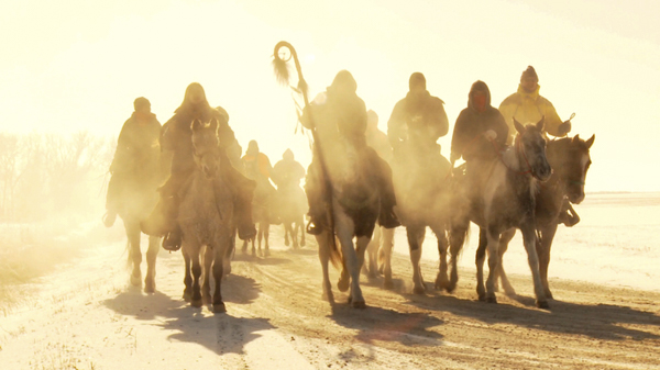 Eine Gruppe von Reitern reiten auf ihren Pferden auf einer schneebedeckten Straße, frontal auf die Betrachterin zu. Gegenlicht und Nebel sorgen für eine mystische Stimmung.