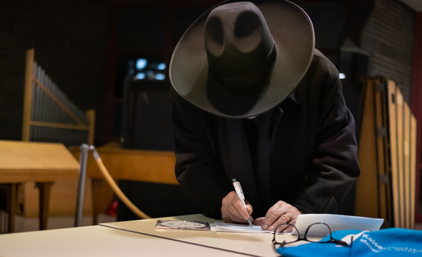 Oskar Gottlieb Blarr beugt sich über den Tisch. Design Hut verdeckt das Gesicht. Er schreibt auf weißes Papier. Vor ihm liegt die Brille. Im Hintergrund steht eine kleine Orgel.