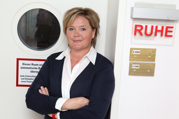 Katja Ruppenthal steht an der Studiotür im WDR. Vor einem runden Fenster. Neben der Tür an der Wand hängt ein Schild, auf dem mit roten Lettern steht "Ruhe". Katja Ruppenthal trägt ein weißes Hemd und hält die Arme verschränkt vor dem Körper. 