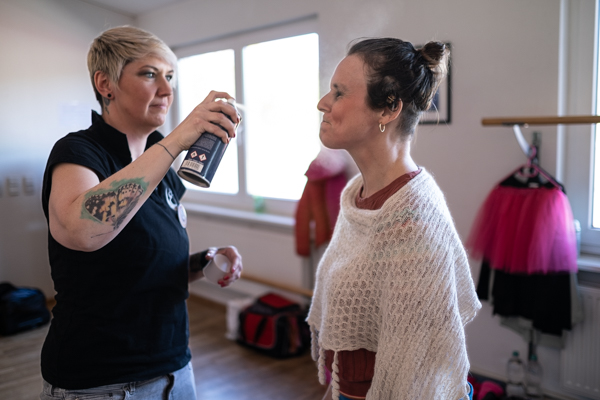 Zwei Frauen stehen im Profil. Davina Reuter, links im Bild, sprüht mit einer Flasche Fixierer ins Gesicht von Alexandra Fröhlich. 